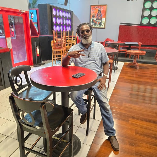 A man with a beard and sunglasses sits at a red table in a restaurant, making a peace sign.