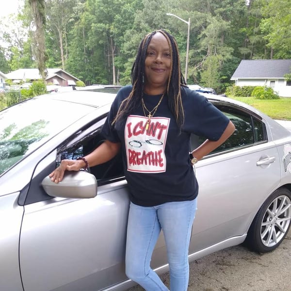 A woman in a black "I Can't Breathe" t-shirt and jeans stands next to a silver car.