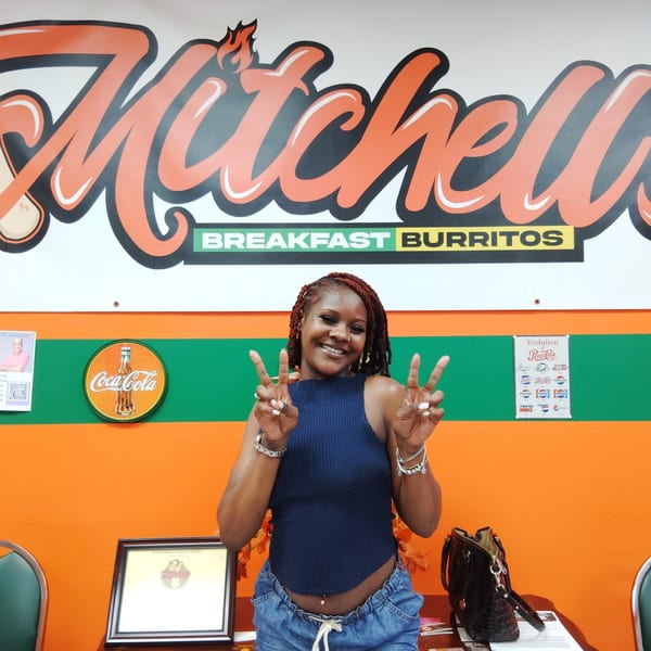 A smiling woman making peace signs in front of the Mitchell's Breakfast Burritos sign.