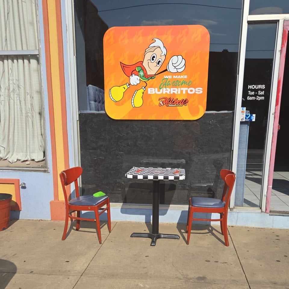Mitchell's Breakfast Burritos storefront with a superhero burrito logo, two red chairs, and a checkerboard table.