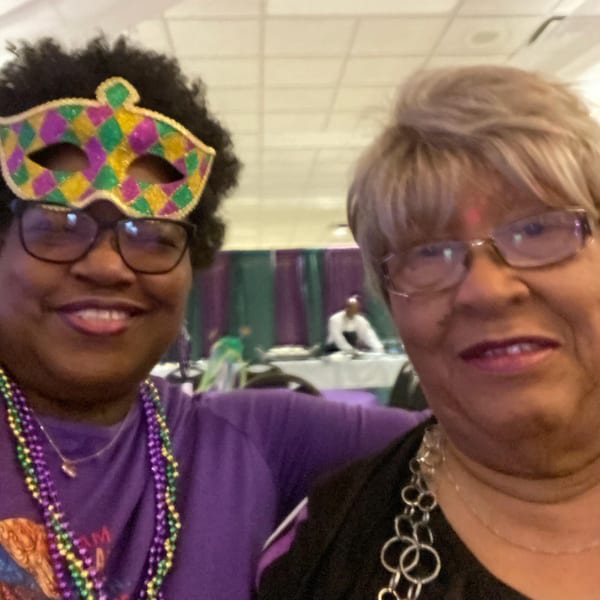 Two women smiling at the camera, one wearing a Mardi Gras mask and beads.