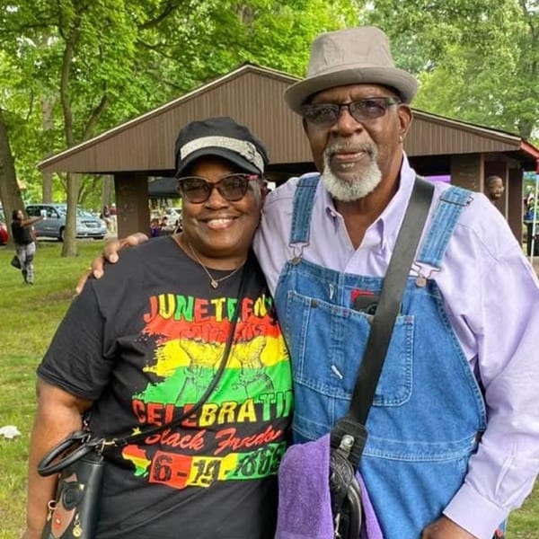 Two people, a woman in a Juneteenth shirt and a man in overalls, smiling outdoors.