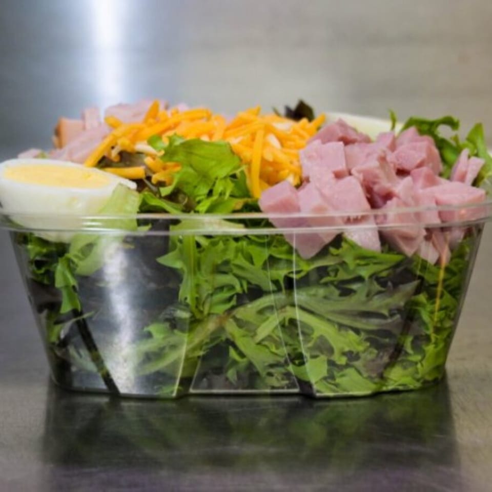 A clear plastic container filled with green salad, diced ham, shredded cheese, and a hard-boiled egg.