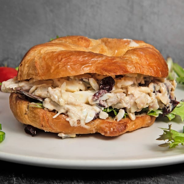 A croissant sandwich filled with creamy chicken salad, lettuce, and dark raisins on a white plate.