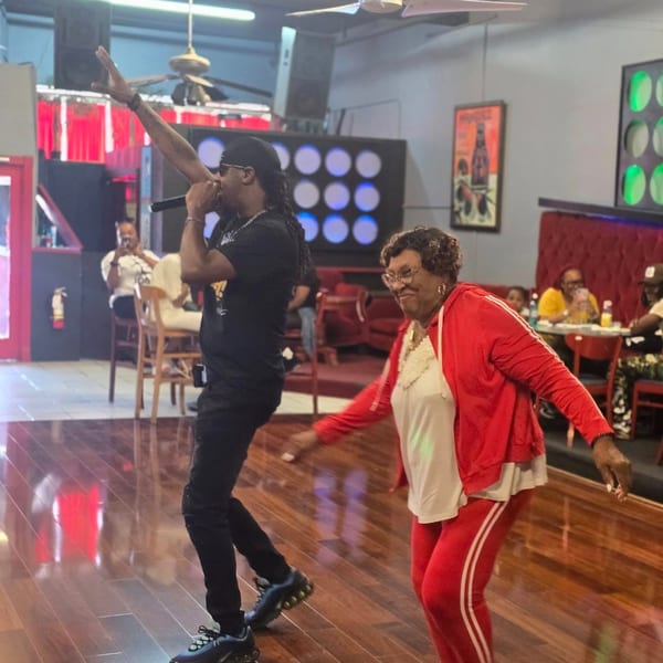A man singing into a microphone and an older woman in red dancing on a wooden floor.